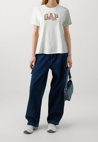 Vit bomulls t-shirt med "GAP"-logotyp och blommönster, i kombination med överdimensionerade mörkblå denimbyxor och vita sneakers, hållande en liten denimväska.