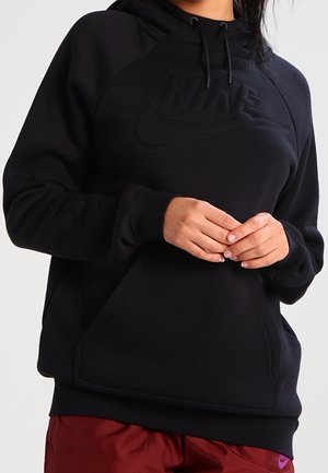 Osoba nosząca czarną bluzę z kapturem Nike z przednią kieszenią i sznurkami, połączoną z bordowymi spodniami z małym fioletowym logo.