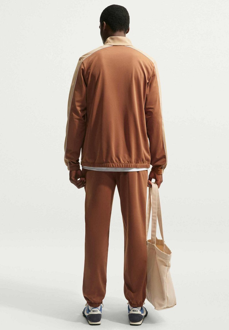 Mand iført brun træningsdragt og hvide sneakers står med ryggen til og holder en beige mulepose på ensfarvet baggrund.