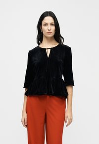Black velvet peplum top with a round neckline and tie front detail, paired with bright red trousers. The fabric has a soft, plush texture.