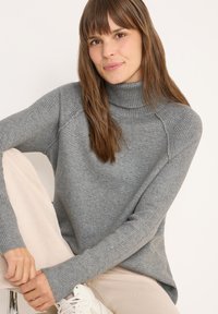 Pull à col roulé en tricot gris avec des poignets et un ourlet côtelés, offrant une coupe décontractée. Texture douce, style décontracté, associé à un pantalon de couleur claire.