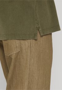 Gros plan de la poche d'un pantalon marron et de l'ourlet d'une chemise vert olive avec une fente latérale, montrant la texture du tissu et les détails des coutures.