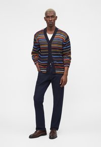 Homme debout, les mains dans les poches, portant un cardigan rayé multicolore, une chemise blanche, un pantalon foncé et des chaussures habillées marron sur fond blanc.