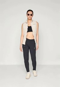 Viegls, bēšas krāsas quilted veste, pieguļošs melns sporta krūšturis, melnas legingas un baltas sporta apavi, papildināti ar saulesbrillēm.