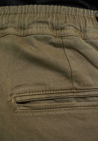 Pantaloni corti di tessuto khaki con vita elastica e una piccola tasca con zip sul retro, caratterizzati da una texture liscia e una leggera lucentezza.