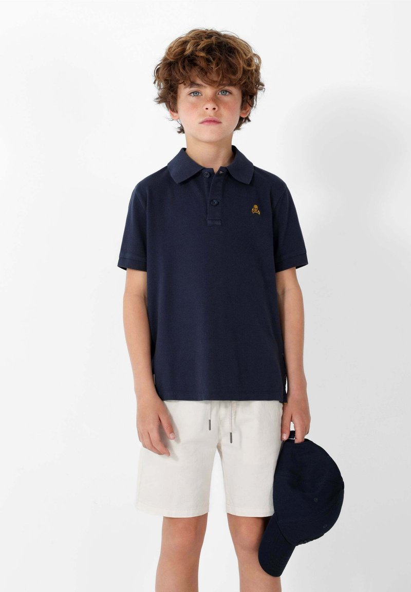 Polo de color azul marino con cuello y mangas cortas, con un pequeño diseño bordado. Combinado con unos pantalones cortos de color claro y una gorra azul marino.