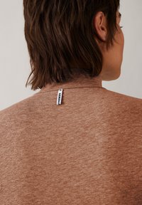 Brauner, weicher Turtleneck aus Stoff mit einer strukturierten Oberfläche und einem kleinen Etikett am Kragen. Das Design ist einfach und figurbetont.