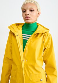 Chaqueta amarilla a prueba de agua con capucha, cremallera y detalles de botones a presión. Dos bolsillos delanteros y textura suave y brillante. Superpuesta sobre una blusa verde.