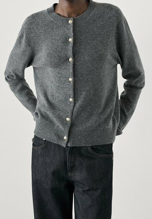 Cardigan en laine grise avec un col rond, arborant des boutons en forme de perles sur le devant, des poignets et un ourlet côtelés. Associé à un jean en denim foncé.