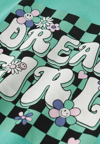 Camiseta gráfica con grandes letras blancas "DREAM GIRL" rodeadas de flores coloridas sobre un fondo de tablero de ajedrez verde azulado y negro.