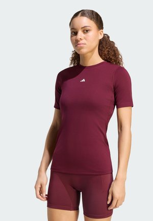 Donna con capelli ricci raccolti in una coda, che indossa una maglietta aderente bordeaux a maniche corte e pantaloncini abbinati su uno sfondo chiaro.
