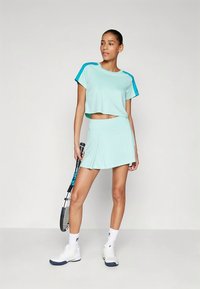 Hele munakasma rohelise tooniga sportlik lühikeste varrukatega ja lühike pealiskleit. Sobitatud volditud munakasma rohelise seelikuga. Valged spordijalatsid ja tennise reket.