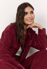 Maroon corduroy jacket with two chest pockets, button closure, and matching pants. Smooth texture, relaxed fit, model leaning with hand on chin.