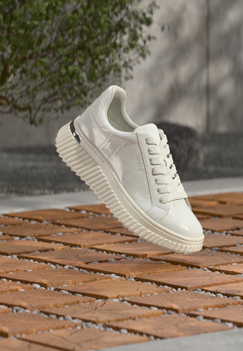 Λευκό αθλητικό παπούτσι με γυαλιστερό, δέρμα με σχέδιο κροκόδειλου, παχιά ραβδωτή σόλα και ασημί λεπτομέρεια. Δεμένα κορδόνια και ορατή μαλακή εσωτερική επένδυση.