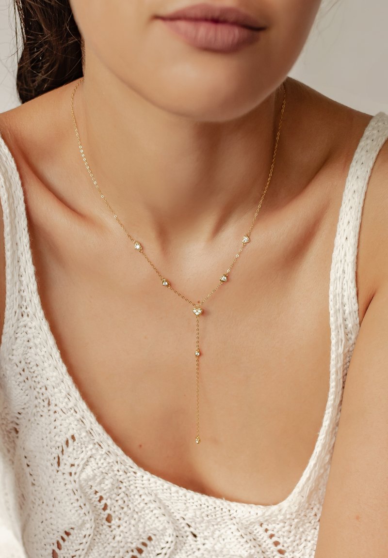 Goldkette mit kleinen, klaren Edelsteinen, mit einem gestuften Design und einem Anhänger; getragen über einem verknoteten weißen Oberteil.