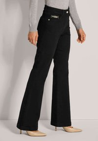 Schwarze Flared-Jeans mit glatter Textur und goldenen Beschlägen am Bund, kombiniert mit beigen spitzen Schuhen.