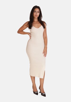 Frau steht mit der Hand in der Hüfte, trägt ein ärmelloses beige geripptes Midikleid und schwarze spitze Absätze mit Schleifendetails.