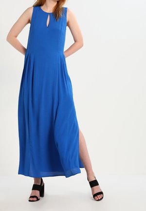 Robe maxi en tissu bleu vif, avec un décolleté en forme de goutte, un design plissé et une fente haute sur le côté. Associée à des sandales à plateforme noires.