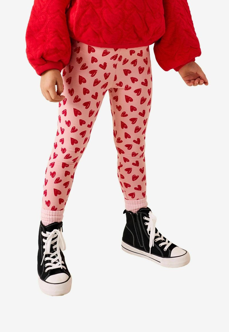 Mallas rosa claro con patrones de corazones rojos, combinadas con un suéter rojo texturizado y zapatillas negras de caña alta con suelas y cordones blancos.