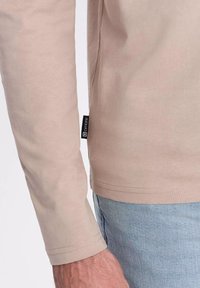 Pitkähihaisen beige-paidan pehmeä rakenne, jossa suora helma ja pieni musta etiketti sivussa. Yhdistetty vaaleansinisiin farkkuihin.