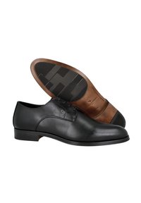 Zapato de vestir de cuero negro con un acabado liso, puntera redondeada, diseño de cordones y una suela de goma marrón en contraste con un agarre texturizado.