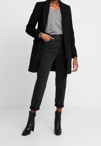 Manteau long noir sur un pull en V gris et un jean noir taille haute, associé à des bottines noires. La tenue allie des lignes épurées et des textures.