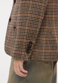 Blazer à carreaux marron avec des accents orange et vert, présentant quatre boutons foncés au poignet, texture lisse et coupe ajustée.