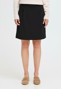 Zwarte knielange rok met geplooide voorkant, elastische tailleband en zijzakken. Draagt met beige instappers en een licht gebreide trui.
