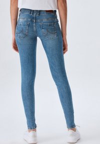 Jeans ajustados de mezclilla de color azul, con una cintura alta, bolsillos traseros con detalles de costura y dobladillos deshilachados. Combinados con zapatillas blancas.