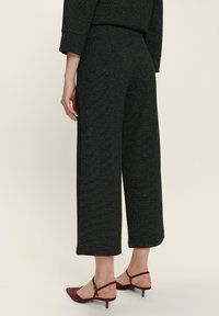 Pantalons croisés noirs en tissu texturé, jambes larges avec une coupe ajustée, associés à des talons slingback bordeaux, présentant une silhouette élégante.