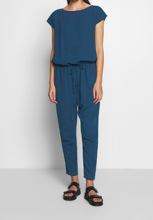 Kobieta w niebieskim kombinezonie bez rękawów z pasem ściągającym w talii i czarnych sandałach na platformie stoi naprzeciwko białej ściany.