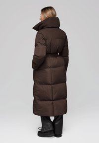 Piumino marrone con collo alto, vita con cintura e motivo trapuntato. Presenta maniche lunghe e lunghezza fino al polpaccio, abbinato a pantaloni neri.