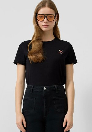 Młoda kobieta o długich brązowych włosach, nosząca duże pomarańczowe okulary przeciwsłoneczne, czarny t-shirt z grafiką koktajlu oraz czarne, wysokie spodnie dżinsowe.