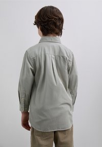 Jasnoszara koszula z długim rękawem, kołnierzykiem, plisą z tyłu i zapinanymi na guziki mankietami. W zestawie z beżowymi, sztruksowymi spodniami.