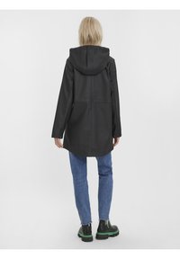 Svart huva regnrock med en slät yta, midilängd och en lätt A-linjeform. Bärs över blå jeans och svarta ankelstövlar med gröna detaljer.