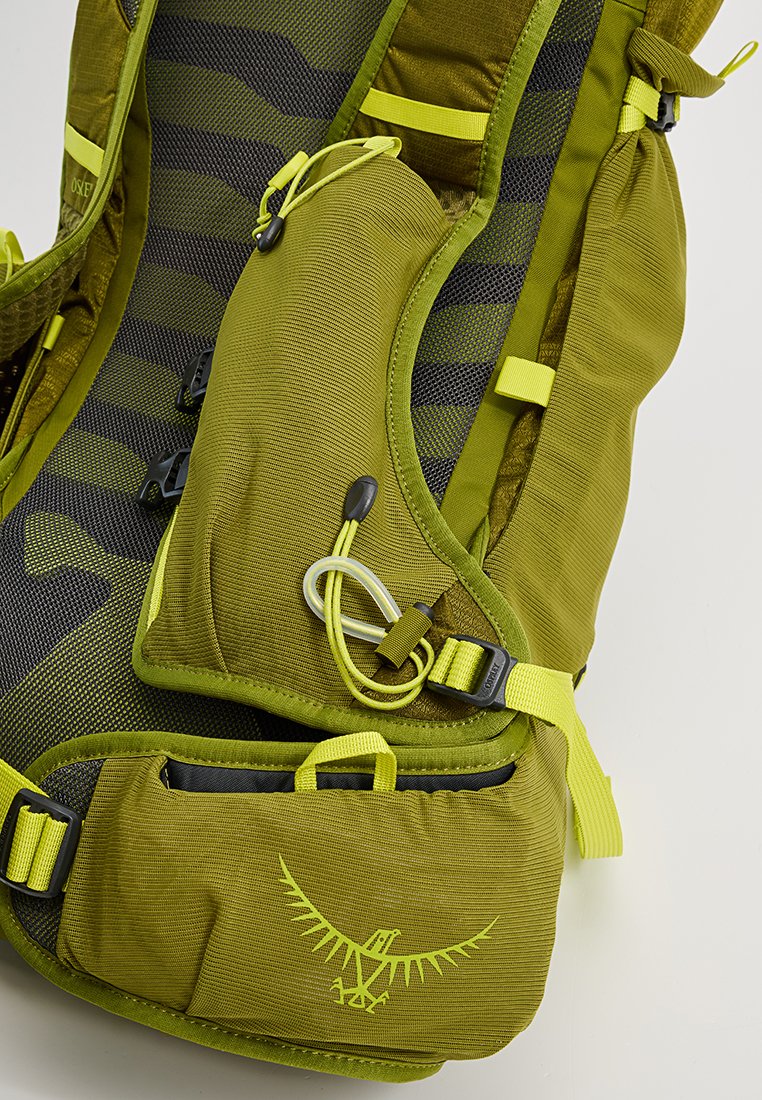 Mochila verde de múltiples bolsillos con textura de malla, caracterizada por correas y herrajes amarillos. Presenta un logo de águila y componentes ajustables.