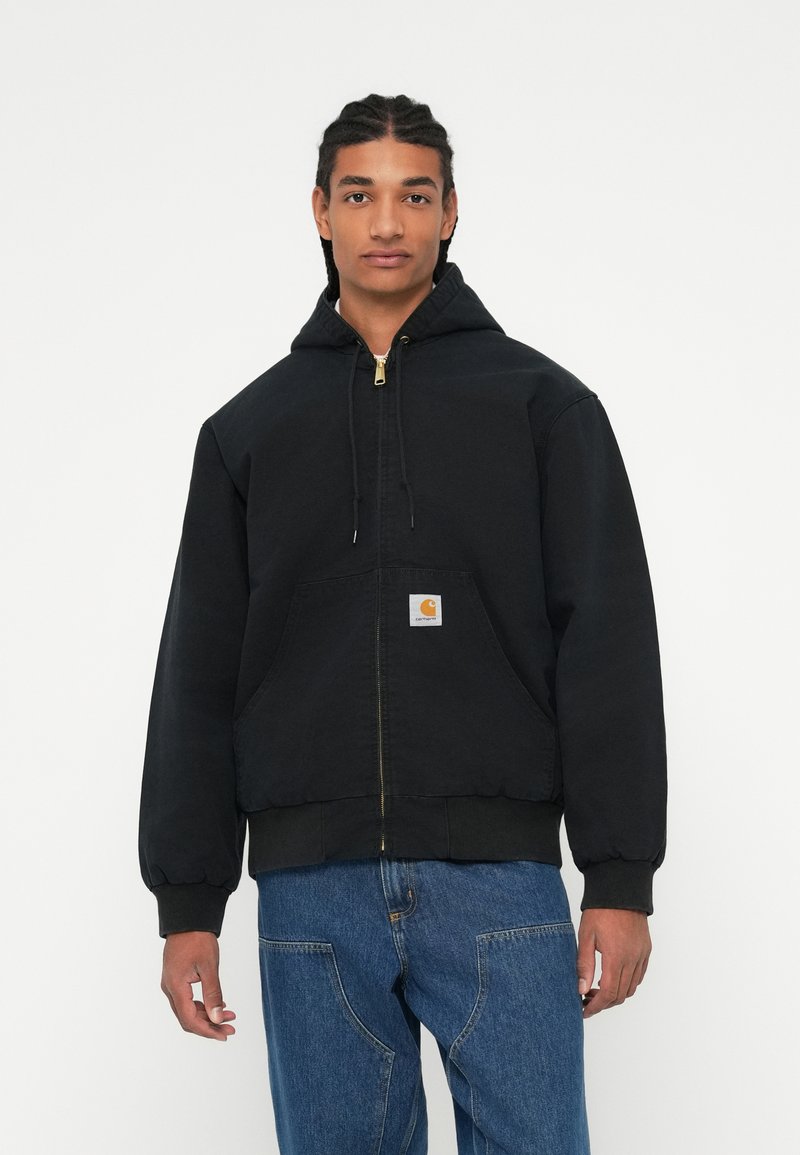 Ung person med fletninger iført en sort hættetrøje med lynlås og frontlomme samt blå jeans, stående mod en ensfarvet hvid baggrund.