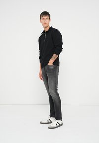 Črna zip-up pulover majica z dolgimi rokavi, v kombinaciji s sivo oprijetimi kavbojkami in belimi športnimi copati, ki imajo črne črte in sive detajle.