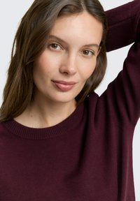 Bordeaux trui met een geribde halslijn, zachte textuur en een relaxed pasvorm. Het model heeft lang, steil haar en steunt met haar hand op haar hoofd.