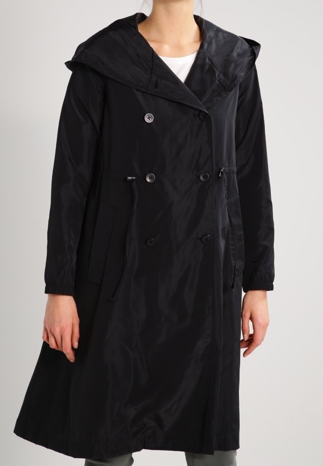 Femme portant un long manteau noir double boutonnage avec un large col et des poches à l’avant, debout devant un fond neutre.