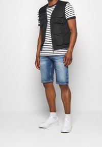 Svart väst med flera fickor över en randig t-shirt. Bärs med denimshorts och vita sneakers. Slät tyg, avslappnad passform.