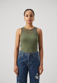 Oliven grønn ermeløs topp med blonde detaljer på skuldrene, kombinert med høy midje, slitte blå jeans. Glatt stoffstruktur.