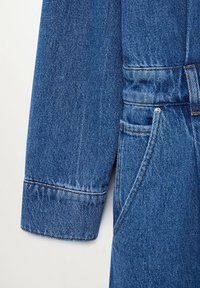 Détail d'une combinaison en denim bleu moyen, avec une coupe ajustée, des coutures visibles, une poche et un col structuré. Tissu doux et texturé.