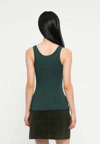 Sieviete ar garām melnām matām, tērpusies tumši zaļā bez piedurknēm topā un tumši zaļā korduroja svārkos, redzama no aizmugures pret vienkāršu fona.