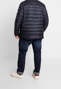 Granatowa kurtka puchowa z pikowanym wzorem, zestawiona z ciemnymi jeansami i białymi trampkami. Kurtka ma miękką, błyszczącą teksturę i swobodny krój.