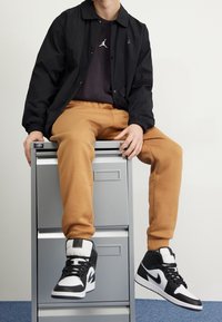 Chaqueta ligera negra con puños elásticos, camiseta oscura con un logotipo, pantalones de chándal color beige y zapatillas altas en blanco y negro.