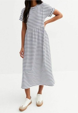 Mujer con vestido midi blanco con rayas horizontales negras y zapatillas blancas con suelas beige y de goma.