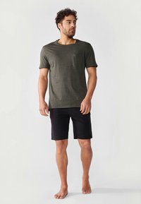 Homme debout pieds nus portant un t-shirt à manches courtes gris foncé avec poche et un short noir jusqu'aux genoux sur un fond uni.