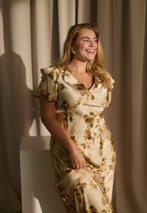Smilende kvinde med langt hår iført en beige blomsterkjole med flæser i ærmerne, der står foran en beige gardinbaggrund.
