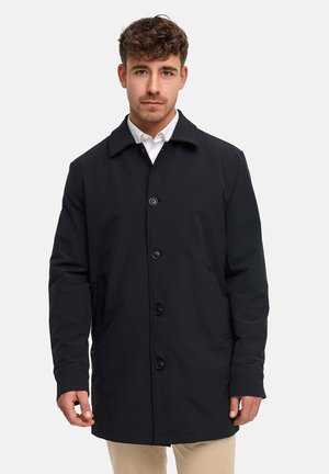 Mann trägt einen schwarzen, geknöpften Mantel über einem weißen Hemd und beigen Hosen und steht vor einem schlichten, hellen Hintergrund.